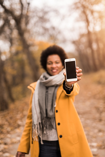 Happy African American woman holding a mobile phone in front of camera while enjoying an autumn day in the park. Focus on foreground.