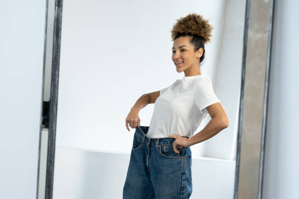 une femme afro-américaine vêtue d’un gros jean avant de perdre du poids, regardant la silhouette devant le miroir, est très heureuse d’avoir réussi à perdre du poids. - weight women medicine lost photos et images de collection