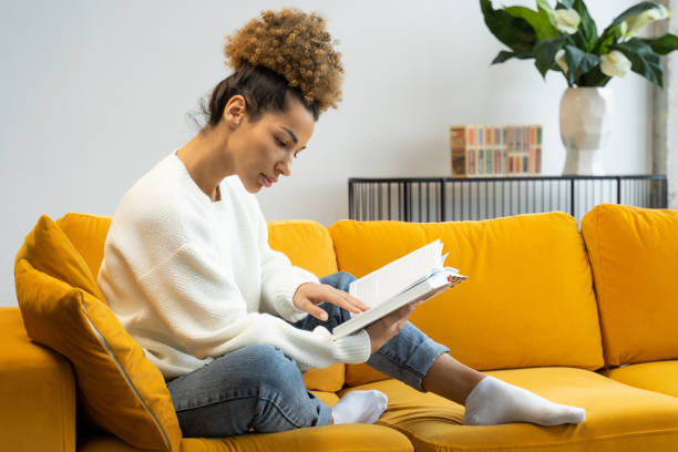atractiva mujer negra de cabello rizado leyendo su novela histórica favorita sentada en un acogedor sofá amarillo - enjoying a novel fotografías e imágenes de stock