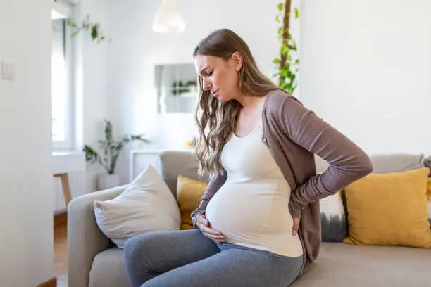 Young pregnant woman suffering from backpain or migraine, feeling sick, sitting on sofa at home, free space. Sad expecting lady touching her big tummy, home alone