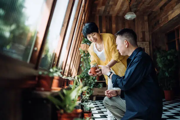 Photo of Happy senior Asian couple spending time together, nurturing houseplants with care at balcony at home. Enjoying life and quality bonding time at cozy home. Retirement lifestyle