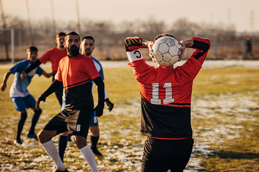 Group of people, male soccer players playing a match on a soccer field on winter day outdoors. One player is throwing in a a ball from out.