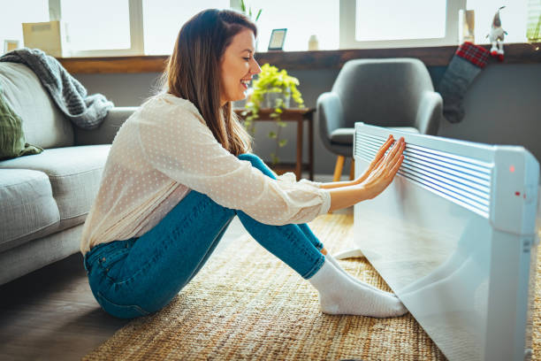 Woman warming hands near electric heater at home. Woman warming hands near electric heater at home. Using heater at home in winter. Woman warming her hands. Heating season. Young woman warming hands near electric heater at home electric heater stock pictures, royalty-free photos & images