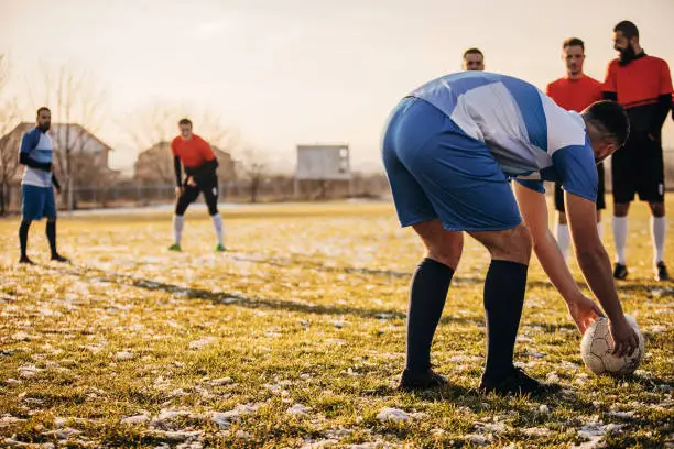 Group of people, male soccer players playing a match on a soccer field on winter day outdoors. One player is preparing a free kick.