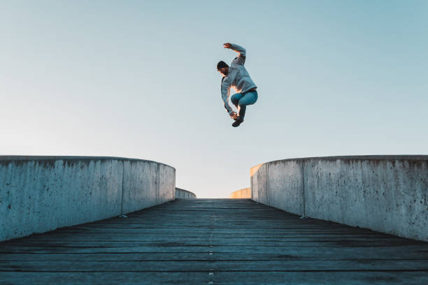 joven caucásico con jeans y sudadera con capucha saltando sobre un puente de concreto. pose de parkour en el aire en el entorno de la ciudad y el cielo despejado - carrera urbana libre fotografías e imágenes de stock