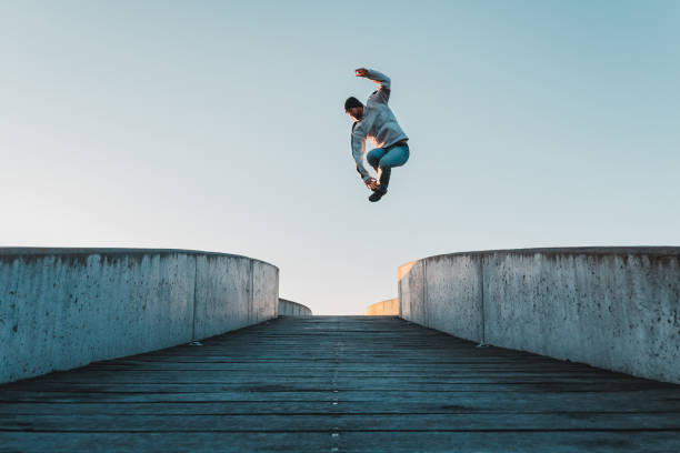 Young caucasian man in jeans and hoodie jumping on concrete bridge. Mid air parkour pose in city environment and clear sky stock photo