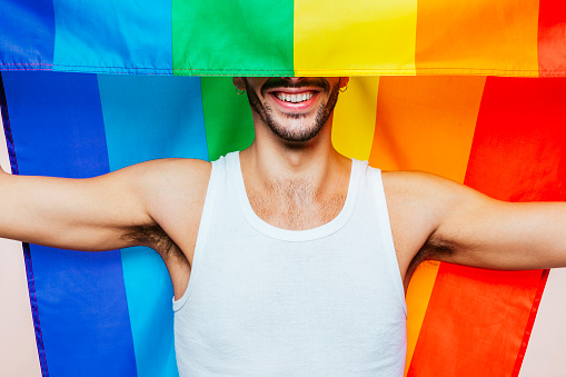 Portrait of cheerful smiling homosexual man with a gay pride flag covers part of\