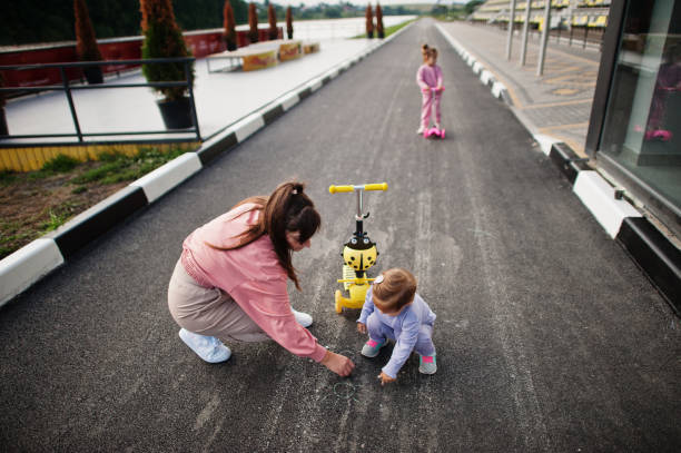 giovane madre elegante con due ragazze all'aperto. la famiglia sportiva trascorre il tempo libero all'aperto con gli scooter. dipinto con gesso sull'asfalto. - 12 23 mesi foto e immagini stock