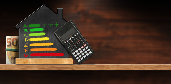 Blackboard in the shape of a house with a chalk drawing of an energy efficiency rating, black calculator and a roll of Euro banknotes, on a wooden table with copy space.