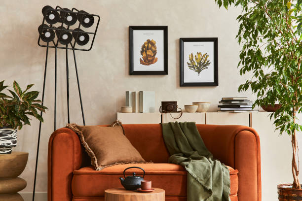 istock Creative composition of stylish living room interior with mock up poster frames, orange sofa, beige commode, coffee table, plant, industrial lamp and stylish personal accessories. Artistic space. Template.