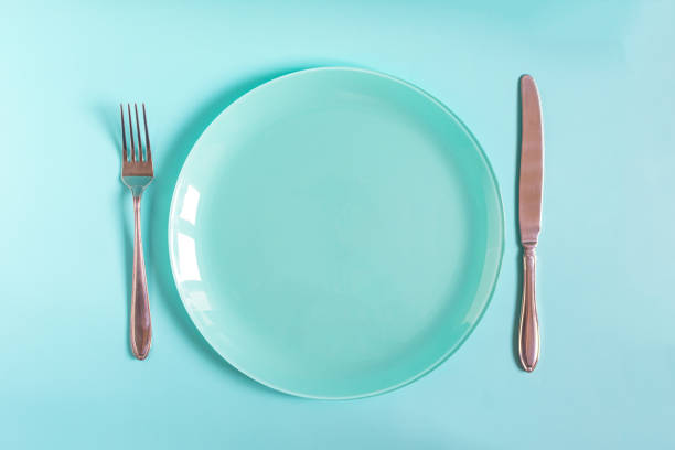 istock Table setting concept: a large blue plate and a fork with a knife on a blue background top view 1360167583