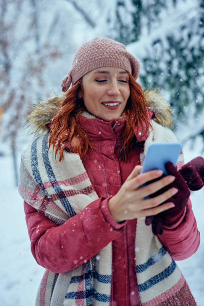 istock Smiling young woman using phone in park at cold winter day 1360172110