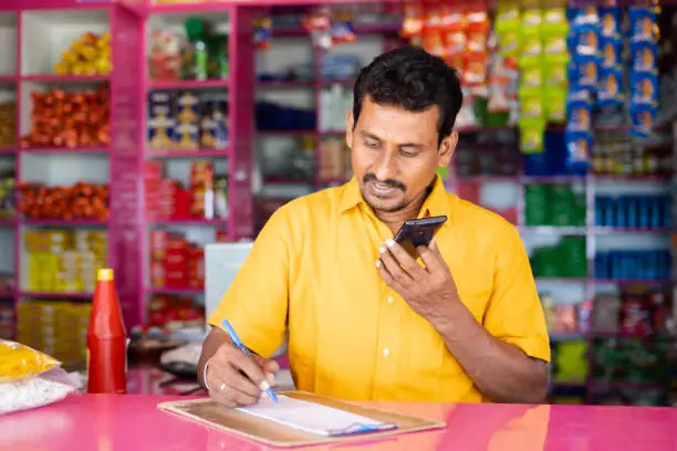 Indian groceries business man noting orders while talking with customer on mobile phone at kirana shop - concept of distance shopping, home delivery service.