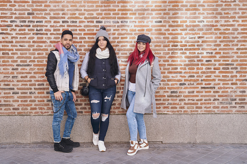 Positive multiracial young male and female friends wearing trendy casual outfits standing near brick wall and smiling at camera