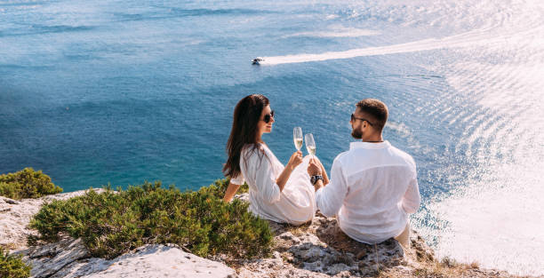 A couple in love drinking champagne on the seashore, panorama. A happy couple on a honeymoon by the sea, panorama. A beautiful couple with champagne on the coast. Honeymoon. Wedding travel. Copy space stock photo