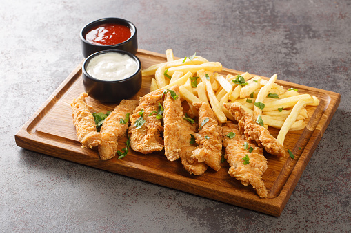 Breaded chicken strips with two kinds of sauces and french fries on a wooden board closeup on a old concrete table. Horizontal