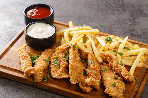 Deep-fried chicken strips served with sauces and fries close-up on a wooden tray on the table. horizontal