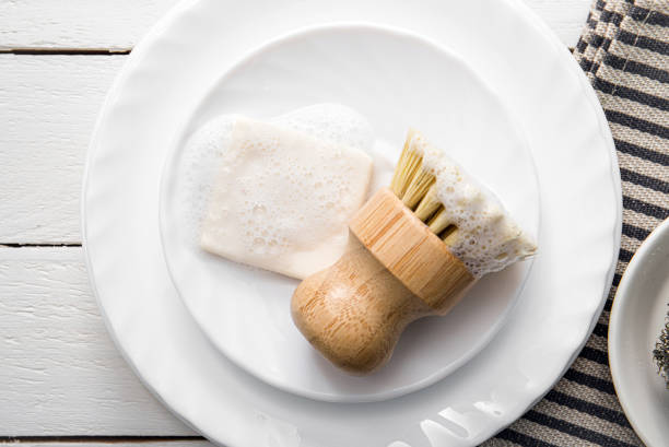 Solid dish washing soap, covered with foam. Zero waste lifestyle. Dishwashing soap on pile of plates with foamy kitchen cleaning sisal bamboo round dish brush. Minimalist white wood background.