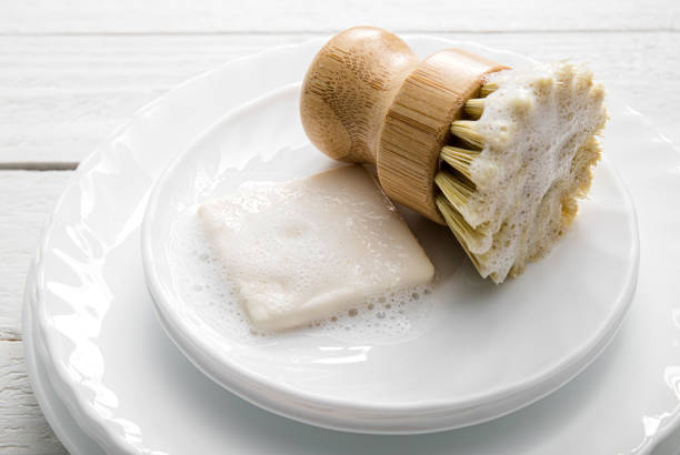 Solid dish washing soap, covered with foam. Zero waste lifestyle. Dishwashing soap on pile of plates with foamy kitchen cleaning sisal bamboo round dish brush. Minimalist white wood background.