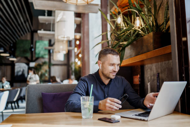 istock Young businessman is working on laptop in restaurant in earpods. Remote work. 1360195521
