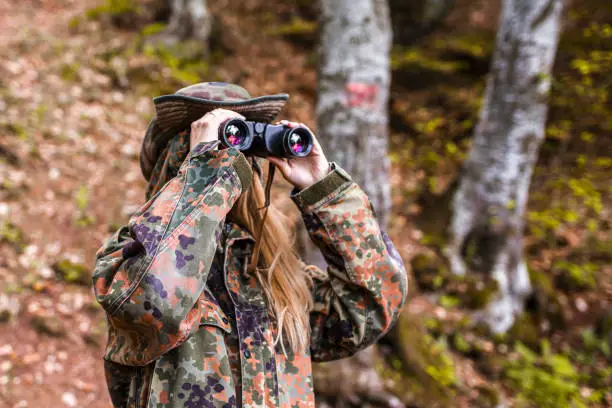 Beautiful woman in camouflage clothing looking through binoculars in the woods