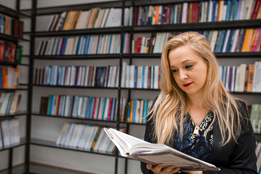 Blonde woman standing in library and reading a book at university