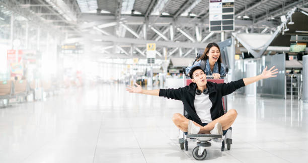 nueva normalidad y concepto de burbuja de viaje.feliz atractivo joven pareja de turistas asiáticos emocionados juntos por el viaje hombre sentado y animando en el carrito de equipaje o carrito de equipaje en el aeropuerto - trolebús fotografías e imágenes de stock