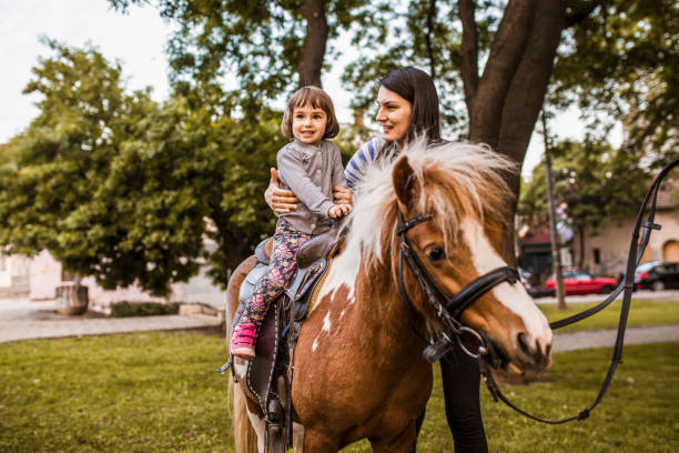 мать держит дочь верхом на пони в парке - pony стоковые фото и изображения