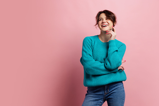 Laughing. Half-length portrait of smiling young beautiful girl in warm knitted sweater isolated on pink color background. Concept of emotions, facial expression, youth, aspiration.