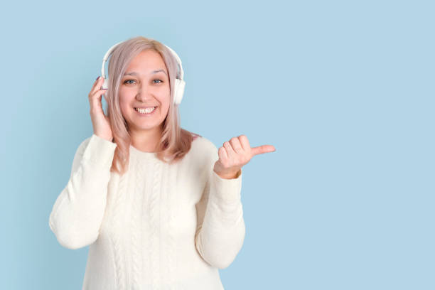una hermosa mujer rubia de 35-40 años escucha música con auriculares inalámbricos y hace un gesto con el dedo hacia arriba mientras está de pie sobre un fondo azul. - 35 40 years audio fotografías e imágenes de stock