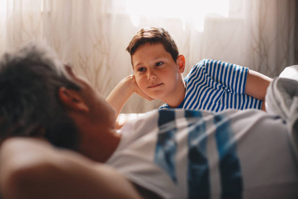 a young boy is talking with his dad. they are lying and relaxing on the bed in the master bedroom. - offspring child lying on back parent imagens e fotografias de stock