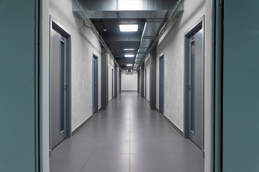 Perspective of a long modern empty office corridor with several gray doors. The floor is tiled and the lights are on.