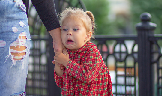 Caucasian little girl of 2 years with scared face holding hand of mother in summertime outdoors