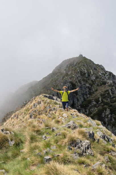 istock Young man stands along grassy mountain ridge, arms wide open 1360224577