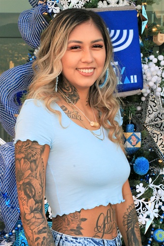 An Indigenous model in front of a decorated Christmas Tree. She is wearing a blue crop top t shirt.
