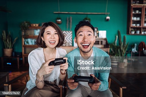 istock Happy young Asian couple sitting on the sofa in the living room, having fun playing video games together at home 1360227632
