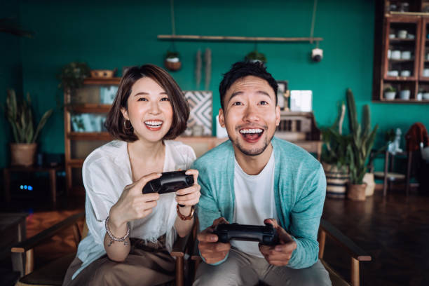Happy young Asian couple sitting on the sofa in the living room, having fun playing video games together at home stock photo