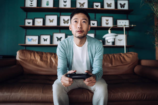 joven asiático concentrado sentado en el sofá de la sala de estar, divirtiéndose jugando videojuegos en casa - espacio masculino fotografías e imágenes de stock