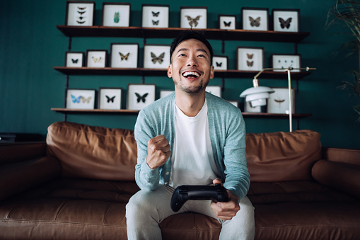Happy young Asian man sitting on the sofa in the living room, shouting in excitement while having fun playing video games at home