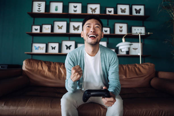 Happy young Asian man sitting on the sofa in the living room, shouting in excitement while having fun playing video games at home Happy young Asian man sitting on the sofa in the living room, shouting in excitement while having fun playing video games at home punching the air stock pictures, royalty-free photos & images