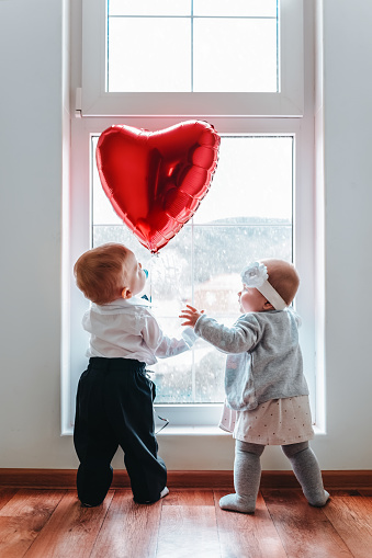 Cute baby boy and baby girl looking at the red heart balloon. Window at the background. The concept of Valentine's day.