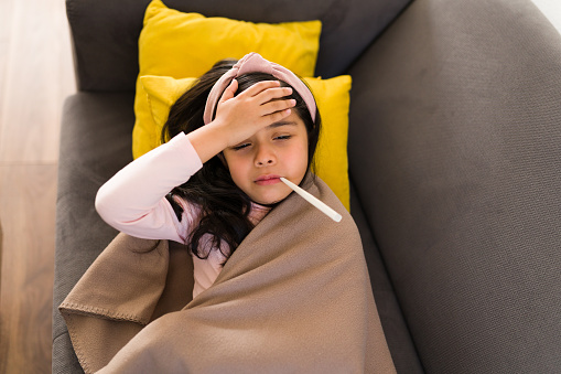 High angle of a latin girl with a thermometer on her mouth. Sick girl putting on hand on her forehead to check her temperature