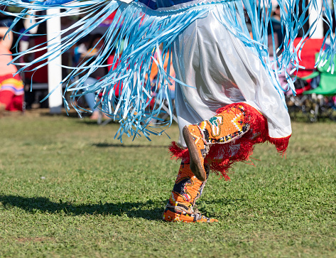 Close up the orange beaded knee high moccasins and shawl decorated with light blue satin ribbons worn by a Native American woman dancing at a pow wow.