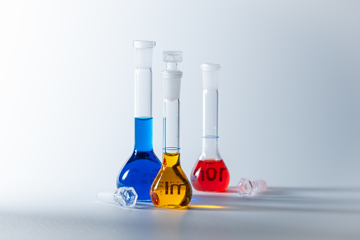 Closeup photo of 3 volumetric flasks on a white background. Each flask filled with different colored solutions, blue, yellow and red.