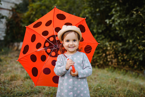 Portrait of a happy smiling little child 2 years old girl holding her red umbrella with polka dots outdoors. Good time after rain.