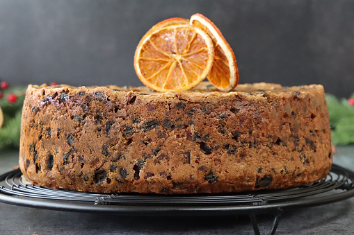 Stock photo showing a close-up view of a homemade Christmas fruitcake with nuts, sultanas and other dried fruits on cooling rack, surrounded by bundles of cinnamon sticks tied with string, star anise, dried citrus orange fruit slices, spruce needles and red berries.