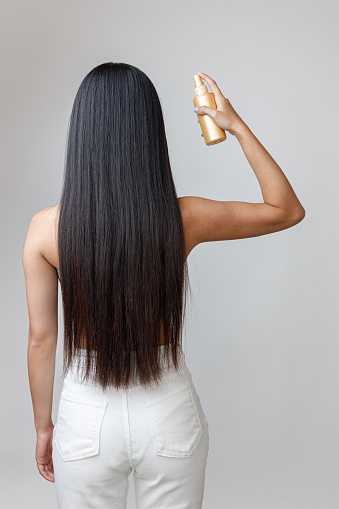 Back view of female person applying hair spray on her long straight hair while standing in studio. Isolated on light gray background