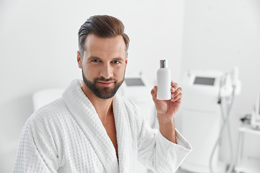 Attractive mature bearded man in white soft bathrobe holds bottle of cosmetic product standing in modern cosmetologycal clinic. Beauty and spa procedures