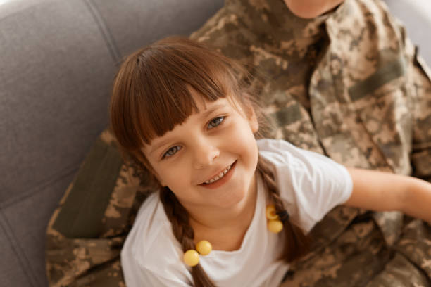 istock Faceless unknown soldier woman wearing camouflage uniform sitting on sofa with charming dark haired female kid with pigtail, kid looking at camera, being glad mother returning from military service. 1360316496