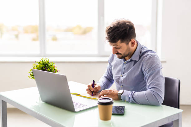 istock Entrepreneur writing notes sitting at his desk. Young man making business plans with papers and computers on his desk in office. 1360337322